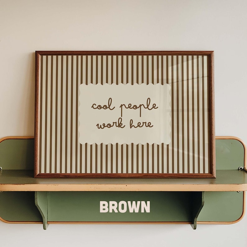 May include: A rectangular framed print featuring a brown and beige vertical striped background with a central scalloped-edge label containing the brown text "cool people work here" displayed in a cursive font style within a dark wood frame.