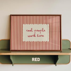 May include: A rectangular framed print featuring a red and cream vertical striped background with a central cream scalloped label containing the red text "cool people work here". The frame is made of dark brown wood and has a simple profile.