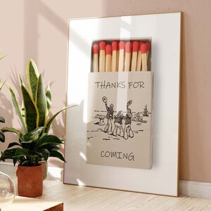 May include: A framed print of a matchbox with the text "Thanks for coming" and a drawing of three people in hats. The matchbox is filled with red-tipped matches.