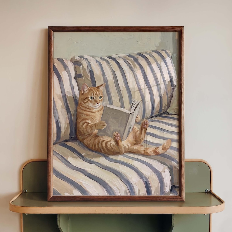 Op de afbeelding: Een ingelijst schilderij toont een rode tijgerkat die een boek leest op een blauw-wit gestreepte bank. Het kunstwerk wordt gepresenteerd in een bruine houten lijst, wat een gezellige en uitnodigende sc&egrave;ne cre&euml;ert.