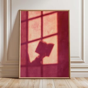 May include: A rectangular wall art print in a light wood frame featuring a deep red and orange gradient background. The image displays a dark silhouette of a person holding an open book, cast against a grid-like window shadow pattern.