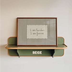 May include: A rectangular framed print with a gray and white vertical striped background and a central white scalloped label featuring the black text I see london I see france. The print rests on a sage green wall shelf with the word BEIGE printed.