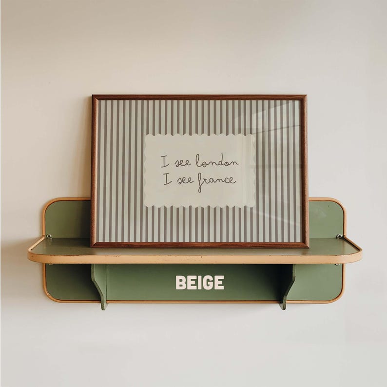 May include: A rectangular framed print with a gray and white vertical striped background and a central white scalloped label featuring the black text I see london I see france. The print rests on a sage green wall shelf with the word BEIGE printed.