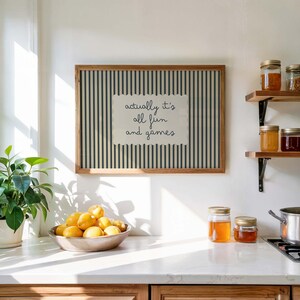 May include: A framed print with the text "actually it's all fun and games" on a striped background. A bowl of lemons sits on a countertop, with jars of preserves on shelves in the background. The frame is made of wood.