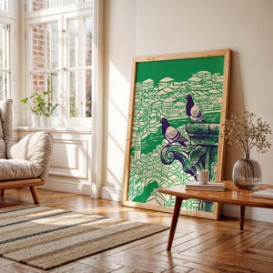 May include: A framed art print featuring a green and white illustration of a cityscape with two pigeons perched on a decorative element. The artwork is displayed in a well-lit room with a wooden table, a rug, and a window.