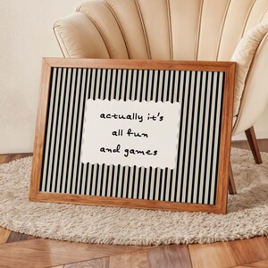 May include: A framed wall art piece with a wooden frame. The artwork features a black and white striped background with a white, scalloped-edged rectangle in the center. The text on the rectangle reads "actually it's all fun and games."
