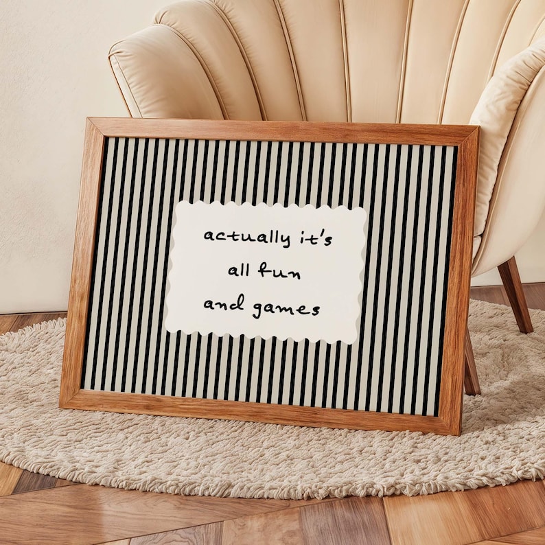 May include: A framed wall art piece with a wooden frame. The artwork features a black and white striped background with a white, scalloped-edged rectangle in the center. The text on the rectangle reads "actually it's all fun and games."