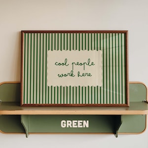 May include: A framed rectangular print featuring a dark green and cream vertical striped background with a central cream scalloped label containing the dark green text "cool people work here". The print rests on a green metal shelf labeled "GREEN".