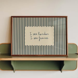 May include: A rectangular wall art print in a dark wood frame featuring a vertical dark blue and cream striped background. In the centre, a cream-coloured scalloped rectangle displays the dark blue handwritten text: I see london I see france.