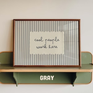 May include: A rectangular framed print with a blue and white vertical striped background and a central scalloped cream label featuring the dark gray text "cool people work here". The frame is dark brown wood, resting on a green metal shelf.
