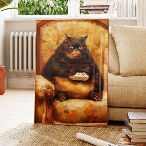 May include: A framed print depicts a large black cat seated in a plush armchair, holding a white video game controller. The cat has a stern expression. The artwork is in a warm, painterly style, with a focus on the cat and its surroundings.