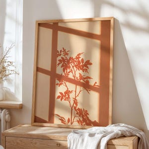 Puede incluir: Una impresión de arte mural rectangular en un marco de madera clara que presenta la silueta de color terracota de una rama con hojas, superpuesta con la sombra geométrica y nítida de un marco de ventana sobre un fondo beige suave.
