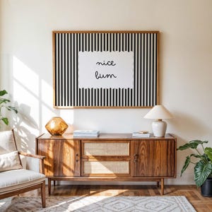 Puede incluir: Un cuadro enmarcado con un fondo a rayas blancas y negras y la frase escrita a mano &laquo; nice bum &raquo;. Sobre un mueble de madera con una l&aacute;mpara y objetos decorativos.