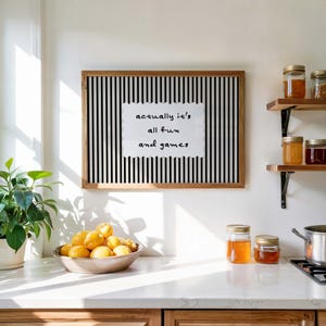 May include: A framed art print with a black and white striped background and the text "actually it's all fun and games". A bowl of lemons sits on a countertop, with jars of preserves on a shelf.