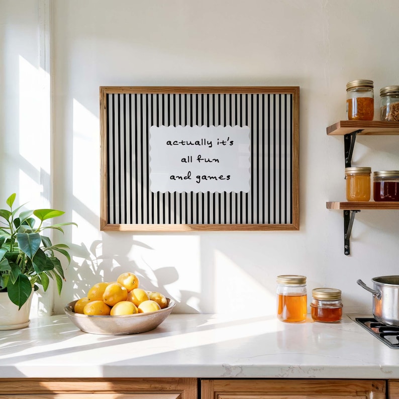 May include: A framed art print with a black and white striped background and the text "actually it's all fun and games". A bowl of lemons sits on a countertop, with jars of preserves on a shelf.