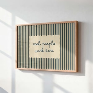 May include: A rectangular wall art piece in a light wood frame featuring a vertical dark blue and beige striped background. Centered is a cream-colored scalloped rectangle with the dark blue text "cool people work here" written in a cursive font.