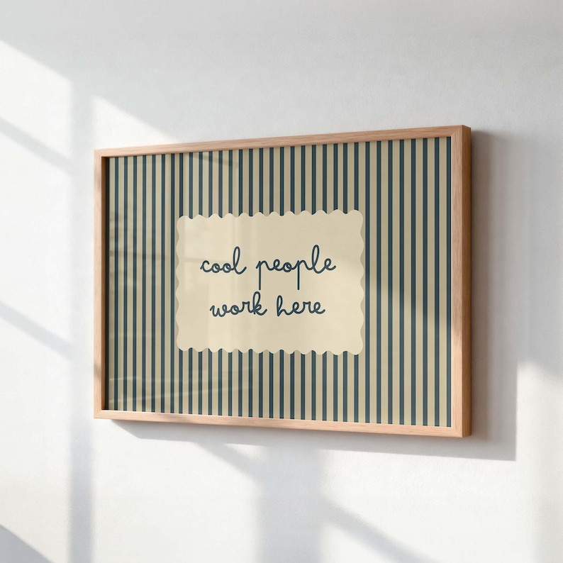 May include: A rectangular wall art piece in a light wood frame featuring a vertical dark blue and beige striped background. Centered is a cream-colored scalloped rectangle with the dark blue text "cool people work here" written in a cursive font.