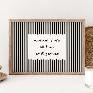 May include: A framed print with a black and white striped background. The print features a white, scalloped-edge rectangle with the words "actually it's all fun and games" in black script. The frame is a light brown color.