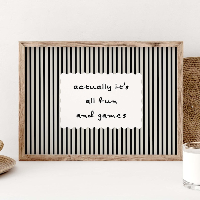 May include: A framed print with a black and white striped background. The print features a white, scalloped-edge rectangle with the words "actually it's all fun and games" in black script. The frame is a light brown color.