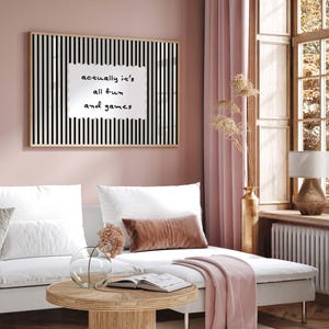 May include: A framed wall art piece with a black and white striped background and the text "actually it's all fun and games" in a white, irregular shape. The art hangs above a white sofa with decorative pillows.