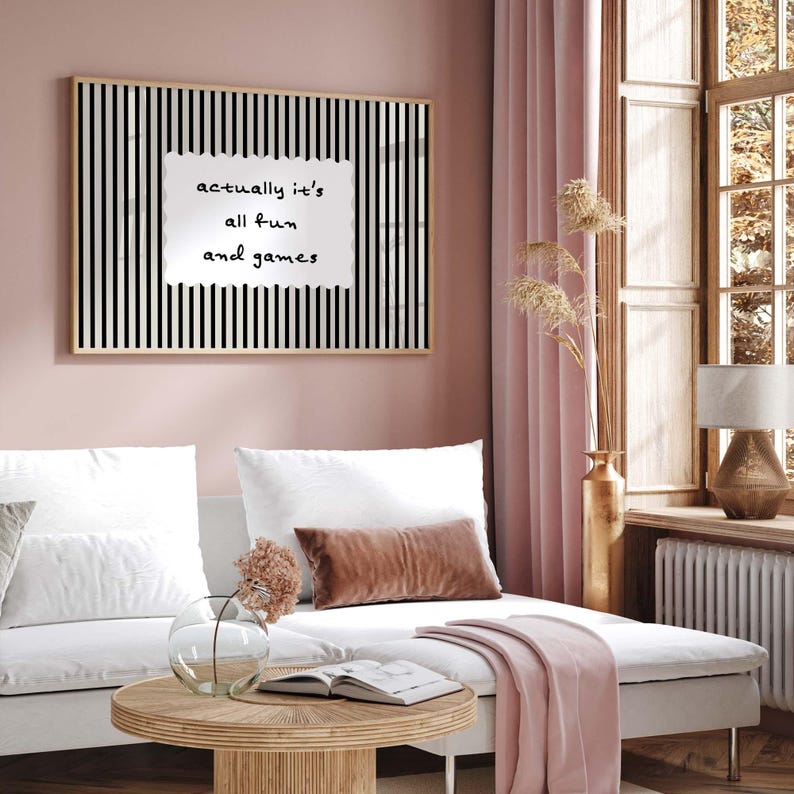 May include: A framed wall art piece with a black and white striped background and the text "actually it's all fun and games" in a white, irregular shape. The art hangs above a white sofa with decorative pillows.