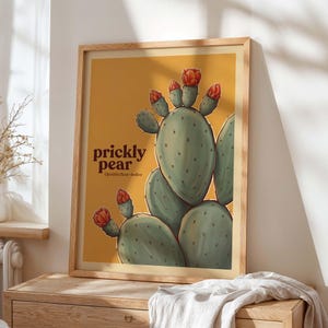 Puede incluir: Una impresión artística enmarcada con una ilustración de un nopal. La obra de arte muestra las palas verdes del cactus y flores rojas sobre un fondo amarillo. Las palabras "prickly pear" están impresas en negrita, en marrón, con el nombre científico debajo.