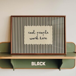 May include: A rectangular framed print featuring a beige background with vertical black stripes and a central scalloped-edge label containing the black text "cool people work here". The frame is dark brown wood, resting on a green metal shelf.