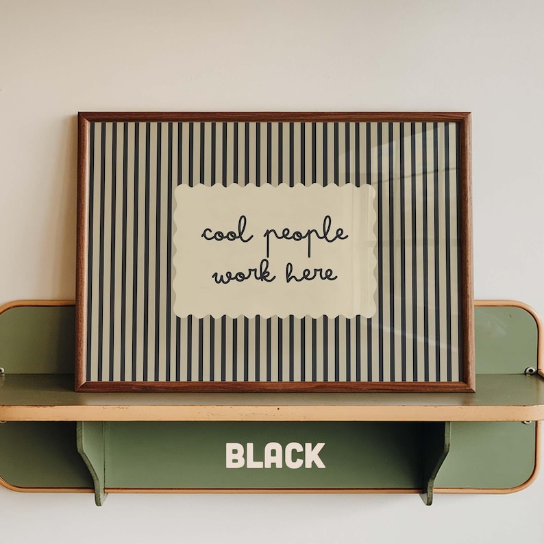 May include: A rectangular framed print featuring a beige background with vertical black stripes and a central scalloped-edge label containing the black text "cool people work here". The frame is dark brown wood, resting on a green metal shelf.
