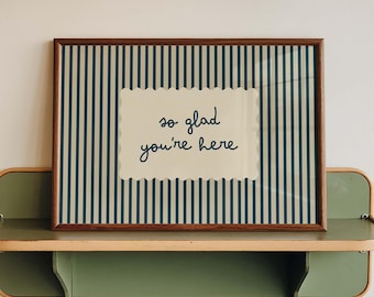 May include: A rectangular framed print featuring a dark blue and cream vertical striped background. In the center, a cream-colored scalloped rectangle displays the dark blue handwritten text "so glad you're here" in a simple, lowercase script font.