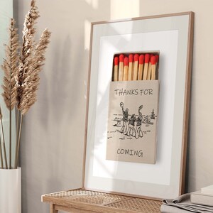 May include: A framed print of a matchbox with red match heads and the text "Thanks for coming". The matchbox features a black and white illustration of three people wearing hats and holding hands.