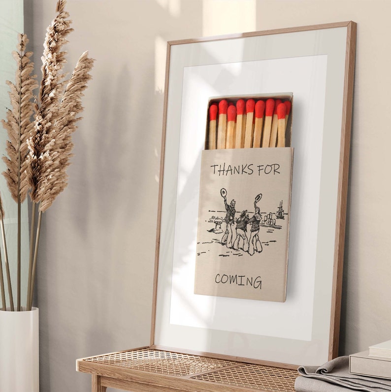 May include: A framed print of a matchbox with red match heads and the text "Thanks for coming". The matchbox features a black and white illustration of three people wearing hats and holding hands.