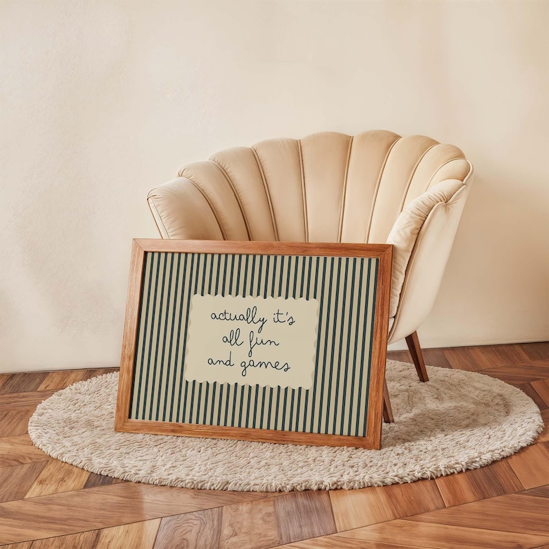 May include: A framed print with a wooden frame, featuring a cream-colored background with vertical dark green stripes. The print includes a cream-colored rectangle with the handwritten text "actually it's all fun and games."