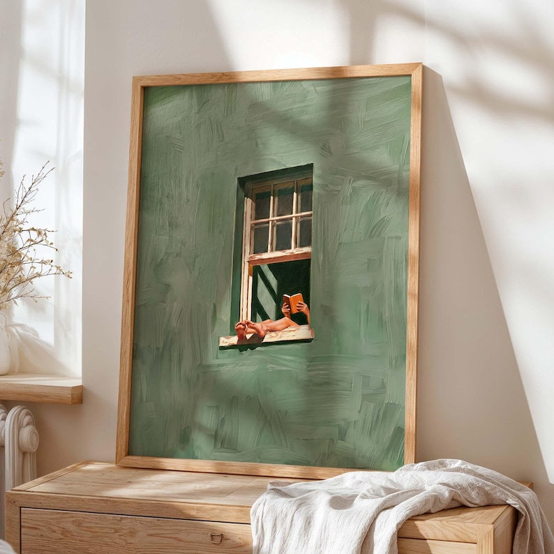 May include: Framed art print featuring a person reading a book by a window. The artwork depicts a green wall with a wooden window frame. The person's legs and hands holding the book are visible. The frame is made of light-colored wood.