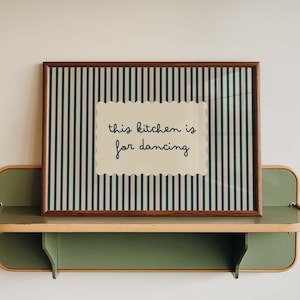 May include: A rectangular wall art print in a dark wood frame featuring a vertical dark blue and cream striped background. Centered is a cream-colored scalloped rectangle with the dark blue text "this kitchen is for dancing" in a cursive font.