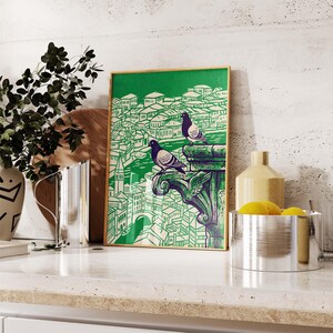 May include: A framed art print featuring a green and white illustration of a cityscape with two pigeons perched on a decorative element. The print has a gold frame and is displayed on a white countertop.