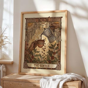 May include: A framed art print featuring a coffee-themed tarot card illustration. The artwork depicts hands pouring coffee into a cup, surrounded by coffee beans and foliage. The text "THE COFFEE" is at the bottom, with a beige background and a wooden frame.