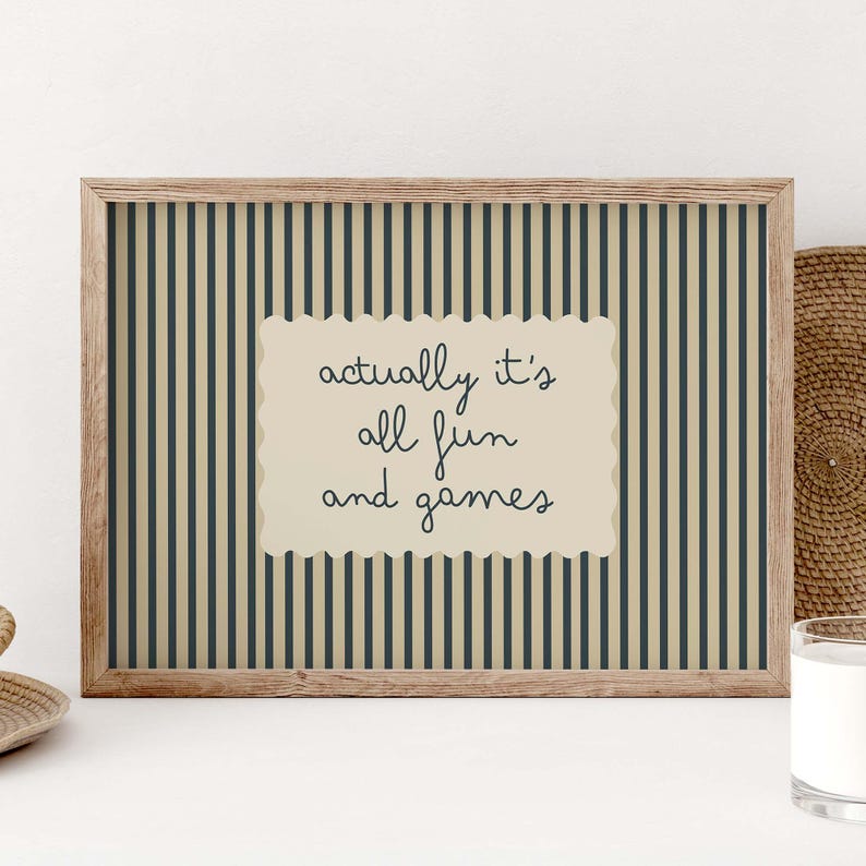 May include: A framed print with a light brown wooden frame. The background has vertical stripes in dark blue and beige. A cream-colored rectangle in the center has the handwritten text "actually it's all fun and games" in dark blue.