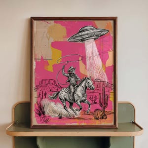 May include: A framed art print featuring a cowboy skeleton riding a horse, roping a UFO. The artwork is set against a vibrant pink and yellow background with desert landscape elements. The print has a wooden frame.