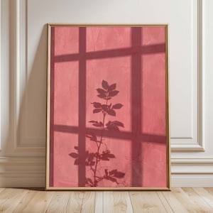 Peut inclure: Une impression d'art mural rectangulaire dans un cadre doré fin avec un fond rose poudré texturé. L'image présente l'ombre sombre et nette d'une grille de fenêtre et la silhouette d'une tige de plante feuillue projetée sur la surface rose.