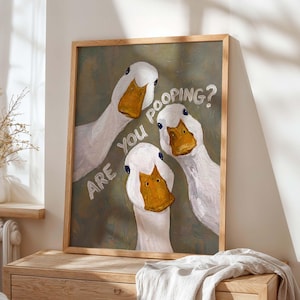 May include: A framed wall art print featuring an oil painting of three white ducks with orange beaks looking forward against a muted olive green background. The text "ARE YOU POOPING?" is written in white capital letters across the center of the image.
