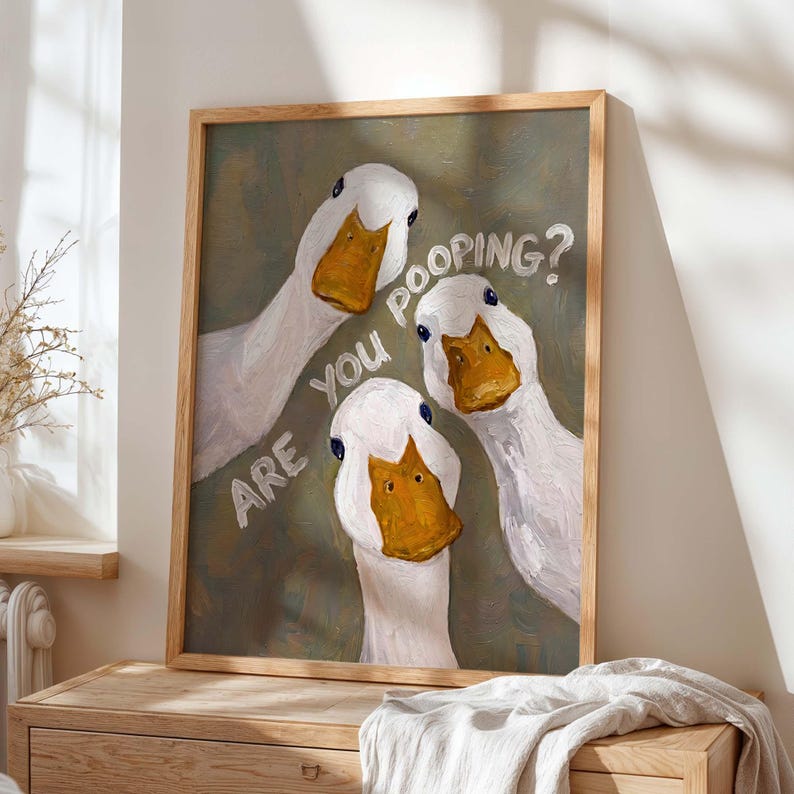 May include: A framed wall art print featuring an oil painting of three white ducks with orange beaks looking forward against a muted olive green background. The text "ARE YOU POOPING?" is written in white capital letters across the center of the image.
