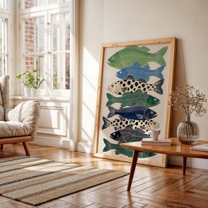 May include: A framed art print featuring a stack of stylized fish in shades of green, blue, and black, with some fish having a spotted pattern. The artwork is displayed in a light-colored wooden frame, leaning against a wall in a well-lit room.