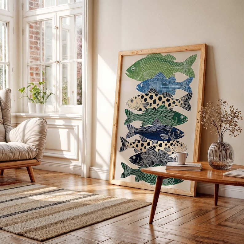 May include: A framed art print featuring a stack of stylized fish in shades of green, blue, and black, with some fish having a spotted pattern. The artwork is displayed in a light-colored wooden frame, leaning against a wall in a well-lit room.