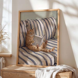 Op de afbeelding: Een ingelijste prent toont een oranje tijgerkat die een boek leest op een blauw-wit gestreepte bank. Het kunstwerk staat in een lichte houten lijst, geplaatst op een houten dressoir. De afbeelding cre&euml;ert een comfortabele, ontspannen sfeer.