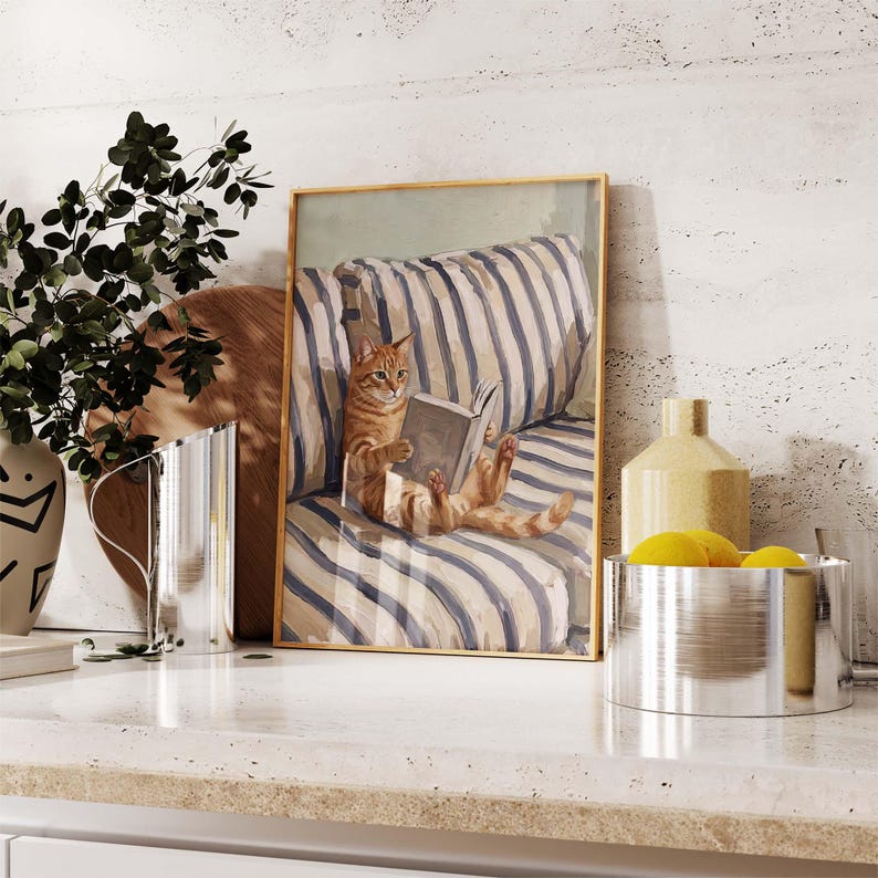 Op de afbeelding: Een ingelijste prent toont een oranje tijgerkat die een boek leest op een blauw-wit gestreept kussen. Het kunstwerk heeft een gouden lijst en wordt tentoongesteld op een wit oppervlak met decoratieve items.