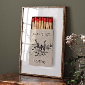May include: A framed print of a matchbox with the text "THANKS FOR COMING" and a drawing of four people in hats. The matchbox is filled with red-tipped matches.