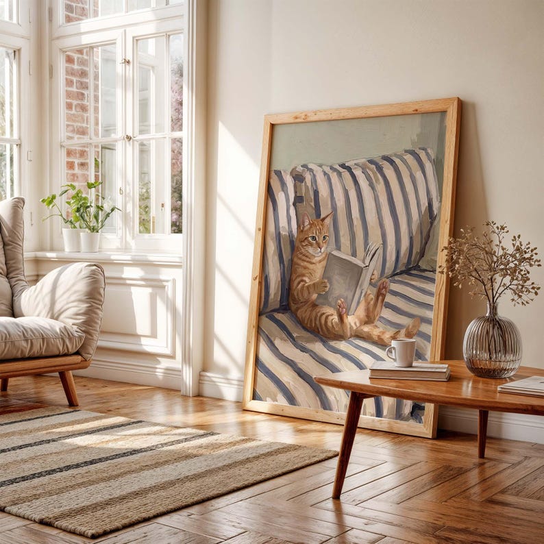 Op de afbeelding: Een ingelijst kunstwerk toont een oranje tijgerkat die een boek leest terwijl hij op een gestreept kussen ligt. Het kunstwerk zit in een lichte houten lijst. Een kleine houten tafel met een koffiekopje en boeken staat voor het kunstwerk.