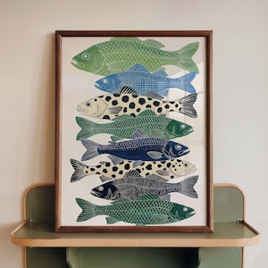 Puede incluir: Una impresión artística enmarcada que presenta ilustraciones de peces estilizados. Los peces están representados en verde, azul y negro, con detalles estampados. La obra de arte se presenta en un marco de madera marrón, ideal para la decoración interior.