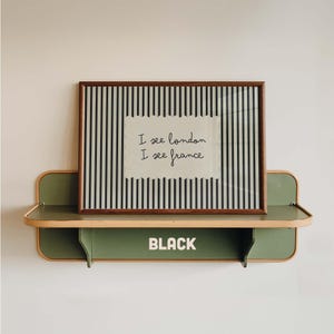 May include: A rectangular framed print with black and white vertical stripes and a central white paper cutout featuring the handwritten text I see london I see france. The frame rests on a sage green wall-mounted shelf with the word BLACK printed.