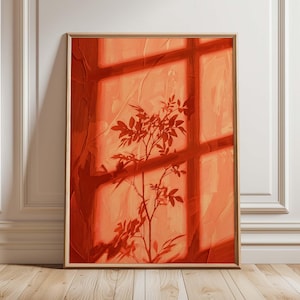 Pode incluir: Uma impressão de arte de parede retangular numa moldura de madeira clara com uma pintura a óleo da silhueta de uma planta projetada numa janela. A obra utiliza uma paleta monocromática de tons laranja quente e terracota com pinceladas.